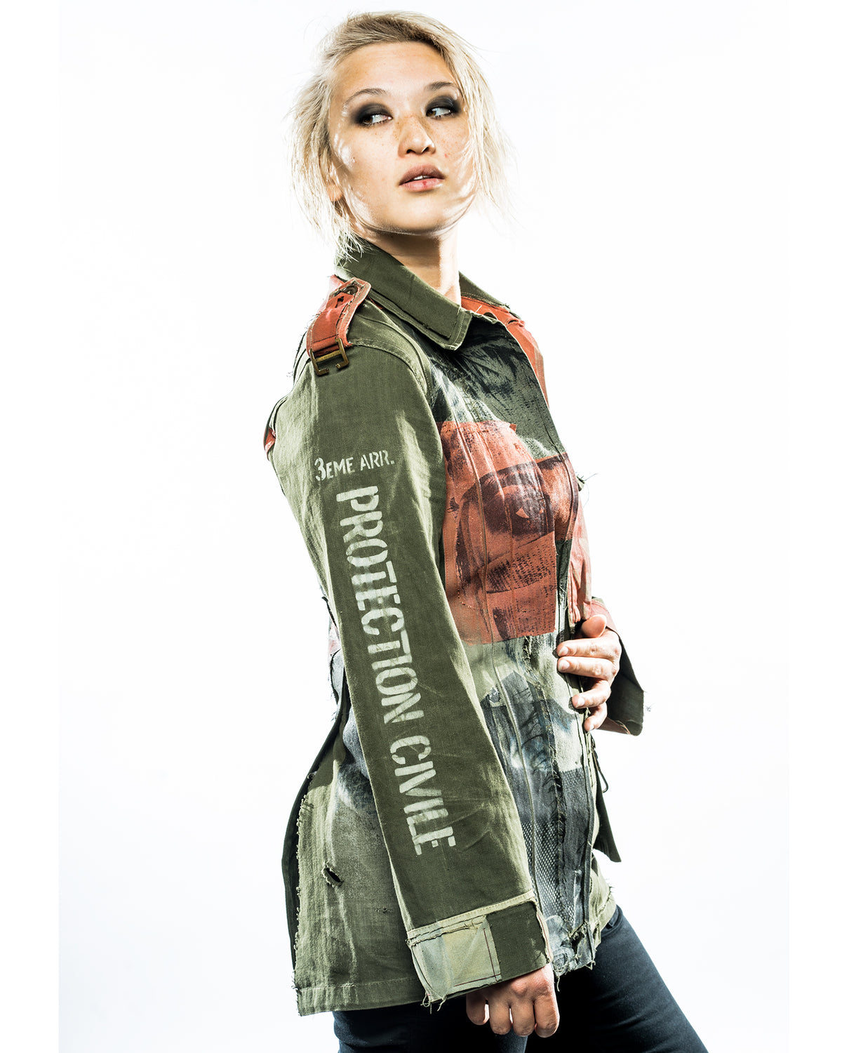 A woman wearing a green deconstructed army jacket with text and graphic embellishments. The jacket features a round neckline, long sleeves, and a front that includes a visible zipper.